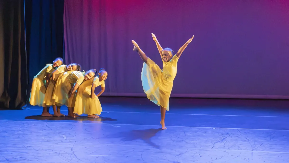 A dancer in flowing yellow holds a high extension as the ensemble gathers behind her.