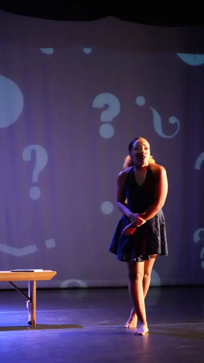A solo performer stands beside a bench against a backdrop filled with projected question marks.