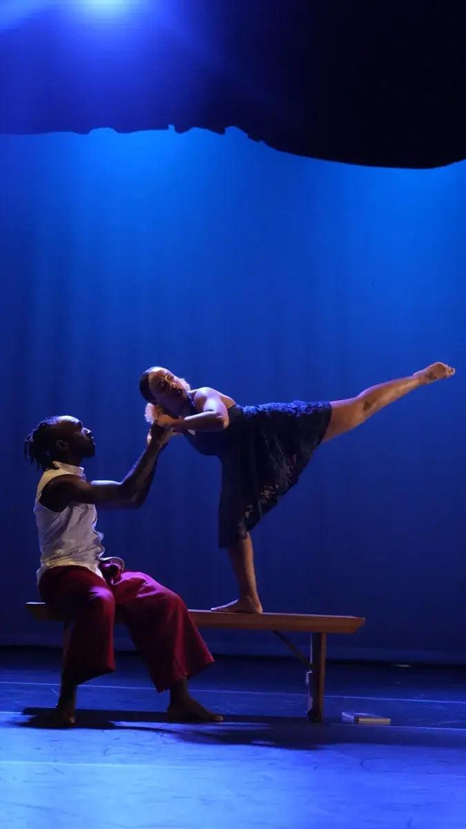 One dancer balances on a bench while another supports her in a blue-lit duet.
