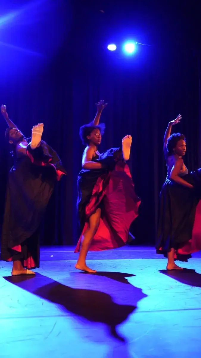 A group of dancers in red and dark costumes perform under vivid blue stage lights.